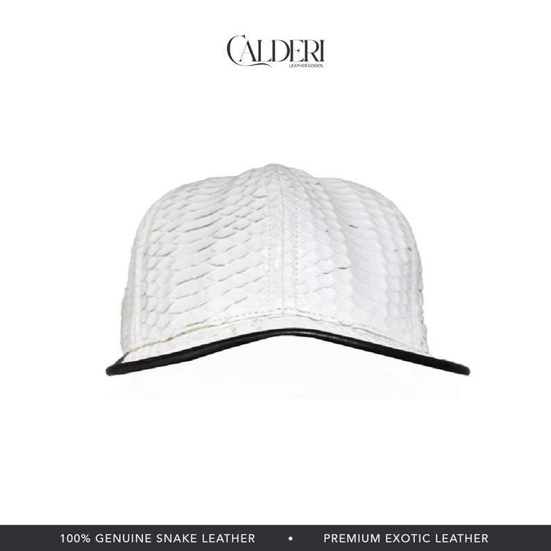 May include: A white baseball cap with a textured pattern, featuring a black brim. The cap is made of genuine snake leather. The brand name "CALDERI" is visible at the top.