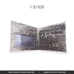 May include: Open bi-fold wallet made of genuine snake leather. The wallet features a snakeskin pattern in shades of gray and white. It has a clear ID window and multiple card slots. The brand name "CALDERI" is displayed at the top.