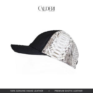 May include: A baseball cap in black and white, crafted from genuine snake leather. The cap has a textured black front and a white and grey snakeskin pattern on the side. The brand name "CALDERI" is displayed.