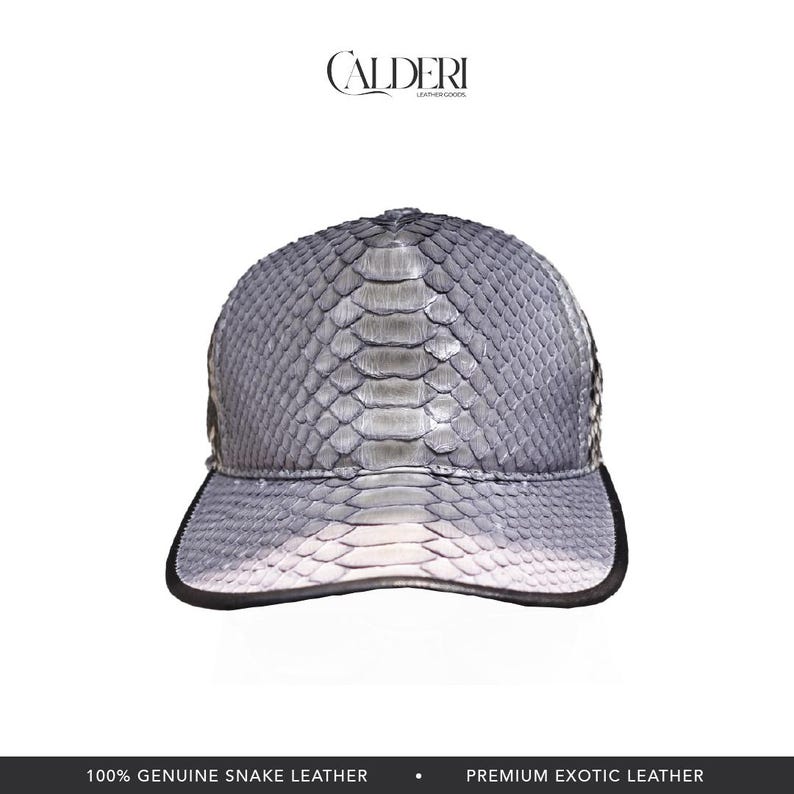 May include: A grey and white snake leather baseball cap with a black brim. The cap is made of genuine snake leather and features the "CALDERI LEATHER GOODS" logo.
