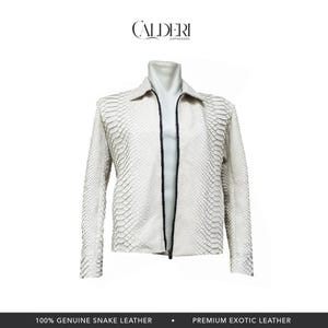 Può includere: Una giacca bianca in pelle di serpente con cerniera e colletto neri. La giacca presenta un motivo strutturato a forma di scaglie. Il marchio "CALDERI" è in alto. Il testo in basso recita "100% GENUINE SNAKE LEATHER" e "PREMIUM EXOTIC LEATHER".