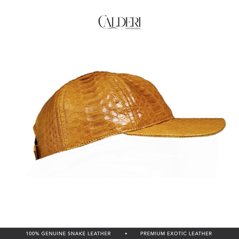 May include: A tan snake leather baseball cap with a curved peak. The cap features a textured pattern and the brand name "CALDERI" at the top. The bottom of the image reads "100% GENUINE SNAKE LEATHER" and "PREMIUM EXOTIC LEATHER."