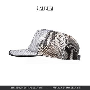 May include: A baseball cap crafted from genuine snake leather. The cap displays a grey and white snakeskin pattern with a black peak. The brand name "CALDERI" is visible at the top.