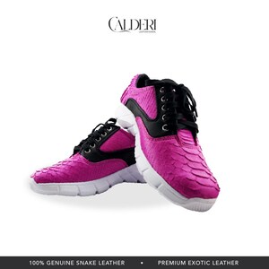 May include: A pair of vibrant pink sneakers with a snakeskin texture. The shoes feature black accents, black laces, and white soles. The brand name "CALDERI" is displayed at the top. The text at the bottom reads "100% GENUINE SNAKE LEATHER" and "PREMIUM EXOTIC LEATHER."