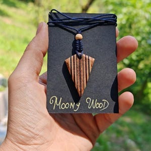 May include: Wooden pendant necklace with a geometric design. The pendant is a light brown color with dark brown stripes and is suspended from a navy blue cord. The text "Moony Wood" is written in gold script on the black card.