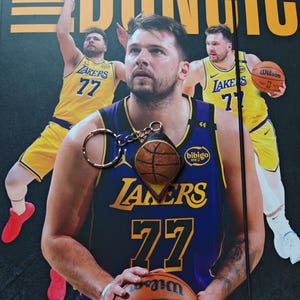 Könnte beinhalten: Ein Basketball-Schlüsselanhänger mit Basketball-Design hängt vor einem Basketballspieler, der ein lila-goldenes Lakers-Trikot mit der Nummer 77 trägt. Der Hintergrund zeigt den Spieler in Aktion mit einem Basketball.