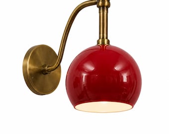 May include: A wall-mounted lamp featuring a glossy red dome-shaped shade and an antique brass finish arm. The fixture includes a circular wall plate, a curved metal support, and a downward-facing light opening with a white interior surface.