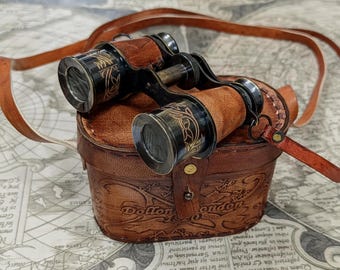 Könnte beinhalten: Ein Fernglas aus schwarzem Messing mit filigranen goldfarbenen Gravuren, das auf einem braunen Lederetui mit passendem Riemen liegt. Das Etui ist auf der Vorderseite mit dem geprägten Schriftzug "Dollond London" versehen.