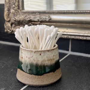 Può includere: Un piccolo contenitore in ceramica fatto a mano pieno di bastoncini di cotone bianchi. Il contenitore presenta una glassa beige screziata, verde scuro e marrone. Il design è semplice e funzionale, adatto per l'uso in bagno o sulla toeletta.