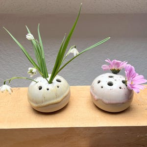 Puede incluir: Dos jarrones de cerámica para flores, cada uno con múltiples agujeros para arreglar los tallos. Uno contiene flores blancas y hojas verdes, el otro flores rosas. Los jarrones son redondos, con un esmalte blanco roto moteado y una banda marrón.