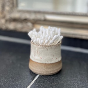 Può includere: Un piccolo contenitore in ceramica fatto a mano con un design bicolore, contenente una collezione di bastoncini di cotone bianchi. La parte superiore è bianco sporco con macchie rossastre, mentre la base è marrone caldo. Il contenitore è su una superficie scura e piastrellata.