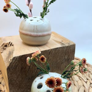 Puede incluir: Dos jarrones de cerámica para capullos, cada uno con múltiples agujeros para tallos. Uno es blanco con una banda rosa, el otro verde pálido. Ambos sostienen flores naranjas y rosas. Los jarrones están sobre un bloque de madera y una estera tejida.