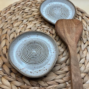 Könnte beinhalten: Zwei kleine, gesprenkelte Keramik-Löffelablagen mit Spiralmuster und ein Holzlöffel. Die Löffelablagen sind hellblau mit braunen Sprenkeln und einem braunen Rand. Der Holzlöffel hat eine mittelbraune Farbe.