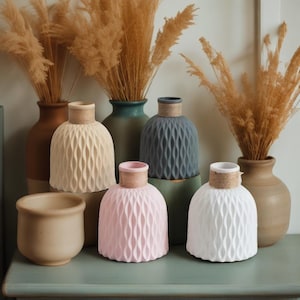 Könnte beinhalten: Eine Sammlung dekorativer Vasen in verschiedenen Farben, darunter Beige, Rosa, Grau und Weiß. Die Vasen haben ein strukturiertes, wellenartiges Muster und sind mit Bindfäden um den Hals verziert. Hohe, getrocknete Gräser sind in mehreren Vasen angeordnet, was eine natürliche, erdige Ästhetik schafft.