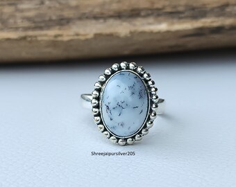 Puede incluir: Un anillo de plata con una piedra preciosa ovalada de color azul claro con vetas grises oscuras. La piedra preciosa está rodeada por un borde plateado con cuentas. El anillo está sobre una superficie clara con un fondo de madera. El texto "Shreejaipursilver205" es visible.