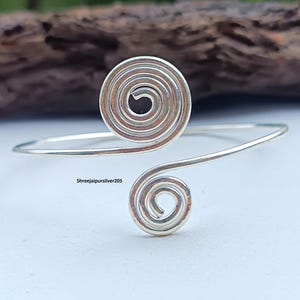Op de afbeelding: Een zilverkleurige manchetarmband met een spiraalvormig ontwerp aan elk uiteinde. De open armband wordt gepresenteerd op een wit oppervlak. De tekst "Shreejaipursilver205" is zichtbaar op de armband. Een delicaat sieraad.