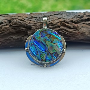 Könnte beinhalten: Ein runder Anhänger mit einem schimmernden Abalone-Muschel-Mittelteil, eingefasst von einem silberfarbenen Metallrahmen und einem blauen Rand. Der Anhänger hat oben eine Schlaufe für eine Kette. Der Text "Shreejaipursilver205" ist unten sichtbar.