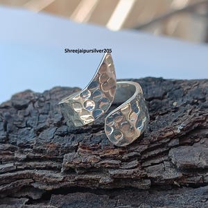 May include: A silver adjustable ring with a hammered texture. The ring has an open design with two curved ends that overlap. The ring is displayed on a piece of dark brown wood. The text "Shreejaipursilver205" is visible.