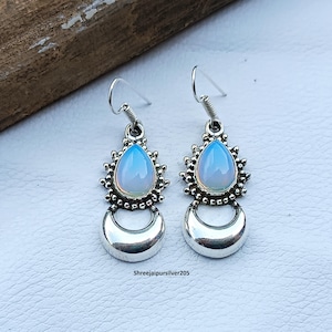 May include: Silver earrings featuring opalescent blue teardrop gemstones. Each gemstone is set in a decorative silver frame, with a crescent moon design below. The earrings are presented on a light surface. The text "Shreejaipursilver205" is visible.