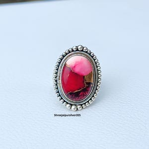 May include: Oval-shaped ring with a vibrant, multi-colored stone. The stone features shades of red, pink, and gold, set in a silver band with a beaded border. The ring's design is eye-catching and detailed, suitable for everyday wear or special occasions.
