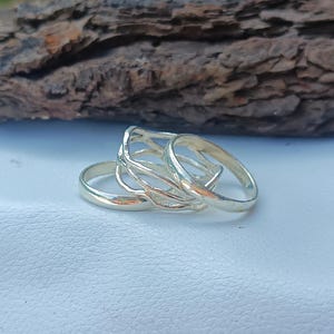 Puede incluir: Una colección de anillos de plata, que incluye un anillo calado texturizado y varias bandas lisas. Los anillos se muestran sobre una superficie blanca con un trozo de madera en el fondo.