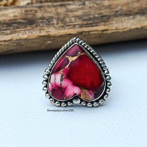 May include: A heart-shaped ring with a silver band and a vibrant stone. The stone features a mix of red, pink, and gold hues. The ring has a beaded border and a twisted rope design around the stone. The text "Shreejaipursilver205" is visible.