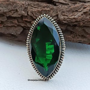 May include: A marquise-cut, emerald-green gemstone ring with a silver-colored, textured band. The gemstone is faceted, reflecting light. The ring is set against a natural wood background.