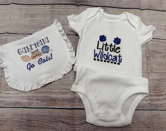 May include: A white baby bodysuit and bib set. The bodysuit has the text "Little Wildcat" in black and blue, with blue flower accents. The bib has the name "Gabriella" and the text "Go Cats!" with a football and cat graphic.