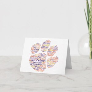 May include: A white greeting card featuring a paw print design made of names in various colors. The text "Mrs. Carlson 2025-2026" is visible. The card is on a white surface with a vase of greenery in the background.
