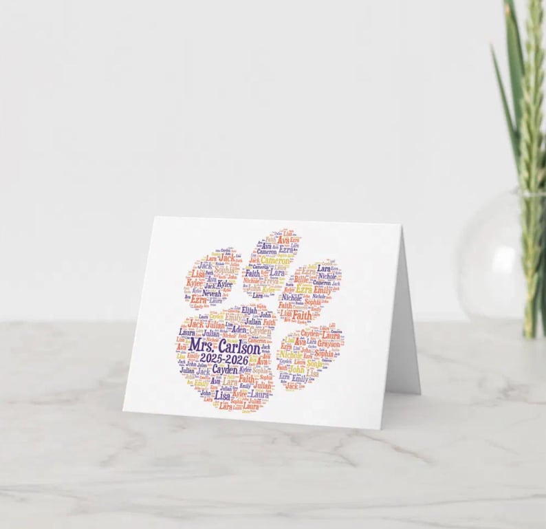 May include: A white greeting card featuring a paw print design made of names in various colors. The text "Mrs. Carlson 2025-2026" is visible. The card is on a white surface with a vase of greenery in the background.