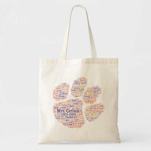 May include: Cream-colored tote bag featuring a paw print design made of names in various colors. The text "Mrs. Carlson 2025-2026" is in the center. The bag has two handles for carrying.