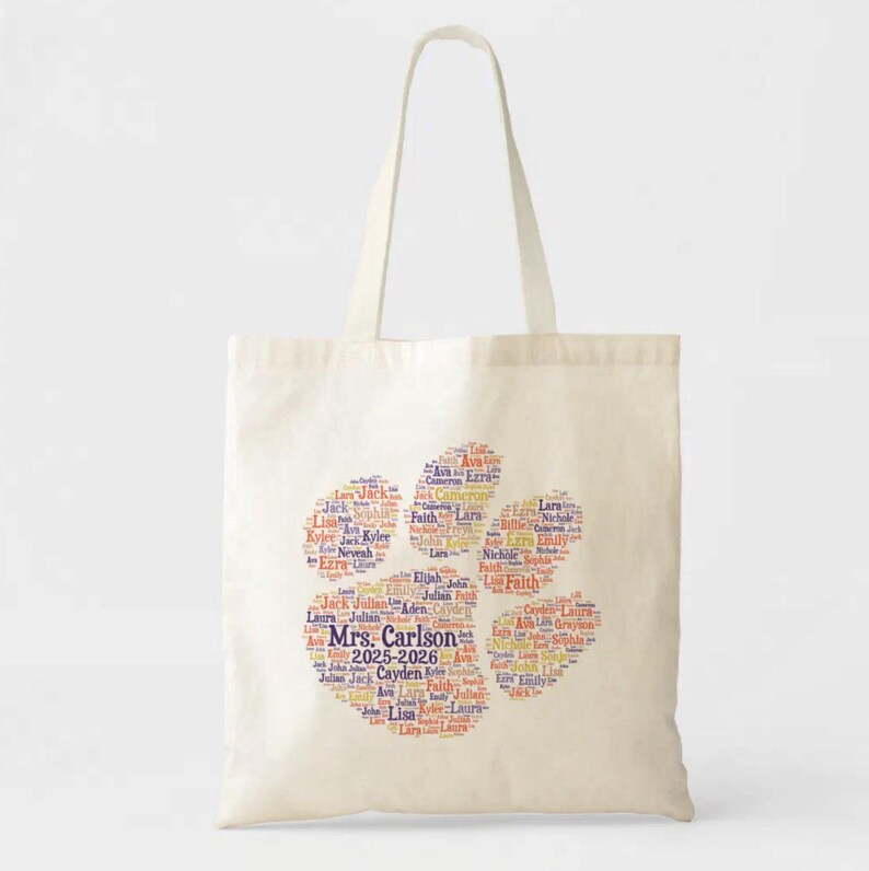 May include: Cream-colored tote bag featuring a paw print design made of names in various colors. The text "Mrs. Carlson 2025-2026" is in the center. The bag has two handles for carrying.