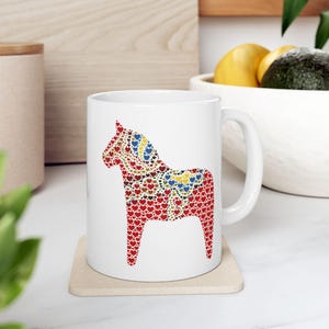 May include: White ceramic mug featuring a colorful Dala horse design. The horse is composed of red, blue, yellow, and brown heart shapes. The mug sits on a beige coaster. A handle is visible on the right side of the mug.