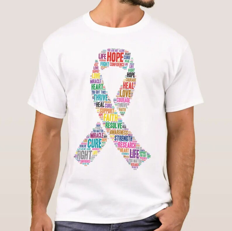 Op de afbeelding: Wit T-shirt met een kleurrijk lintontwerp gemaakt van woorden zoals "hoop", "liefde", "strijd", "genezing", "sterkte" en "onderzoek".