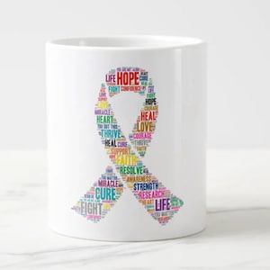Op de afbeelding: Een witte keramische koffiemok met een kleurrijk lintontwerp. Het lint is gemaakt van woorden die te maken hebben met hoop, kracht en genezing, waaronder "Hope", "Heal", "Love", "Fight", "Courage", "Thrive", "Support", "Faith", "Resolve", "Strength", "Research", "Cure" en "Life".