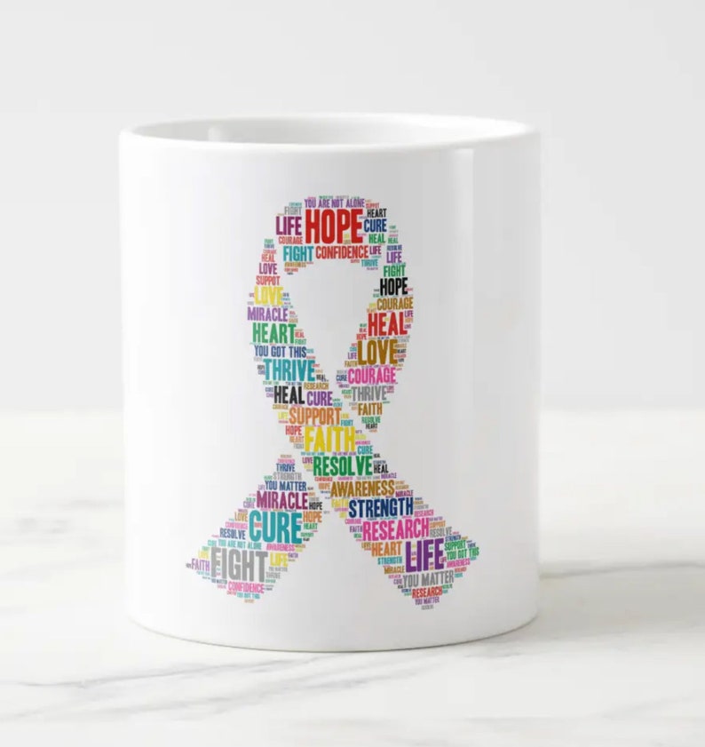 Op de afbeelding: Een witte keramische koffiemok met een kleurrijk lintontwerp. Het lint is gemaakt van woorden die te maken hebben met hoop, kracht en genezing, waaronder "Hope", "Heal", "Love", "Fight", "Courage", "Thrive", "Support", "Faith", "Resolve", "Strength", "Research", "Cure" en "Life".