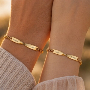 May include: Two gold-colored bracelets with the names "Sophia" and "Liam" engraved on them. The bracelets have a twisted bar design and are connected by a chain. The bracelets are worn on wrists.