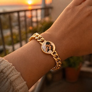 Op de afbeelding: Een goudkleurige kettingarmband met een ronde hanger met een foto van een stel. De armband heeft een dik, getextureerd kettingontwerp. De achtergrond toont een zonsondergang boven water.