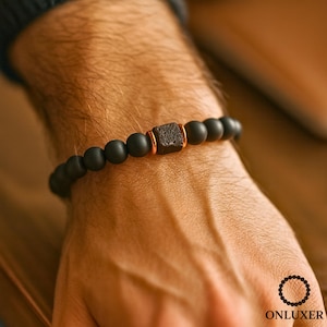 Könnte beinhalten: Schwarzes Perlenarmband mit einem quadratischen, strukturierten, dunkelbraunen Mittelstück und kupferfarbenen Akzenten. Das Armband besteht aus mattschwarzen Perlen und wird an einem Handgelenk gezeigt.