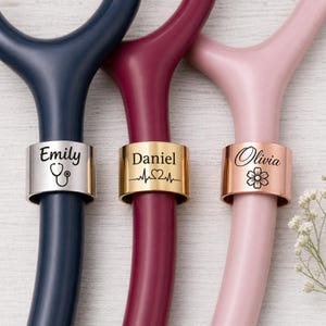 May include: Three personalized stethoscope charms in silver, gold, and rose gold. Each charm is engraved with a name and a symbol: a stethoscope, a heartbeat, and a flower. The stethoscopes are navy, burgundy, and pink.