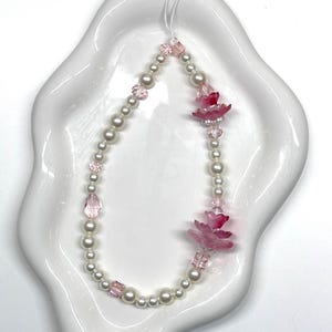 Puede incluir: Un delicado colgante para teléfono con una cadena de perlas de imitación y cuentas rosas. El colgante está adornado con detalles de flores rosas y está suspendido por un cordón blanco. Se muestra en un plato blanco con bordes ondulados.
