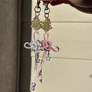 May include: Two handmade keychains with silver-tone hardware. One keychain is white and the other is pink. Each keychain features a star charm and a decorative knot design. The top of each keychain has a floral design with pink and green accents.
