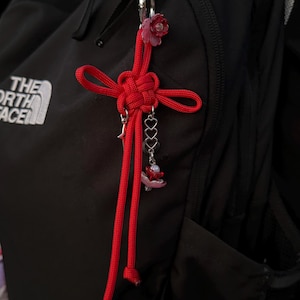 May include: A red decorative knot charm with dangling heart and flower charms, attached to a silver carabiner. The charm is hanging on a black backpack. The charm is a decorative accessory.
