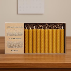 Puede incluir: Una caja con once velas de intención de cera de abeja amarillas. Cada vela tiene un patrón de panal y una mecha blanca. La caja tiene el texto "21 Days of Intention Candles" y "TRSY Gift".