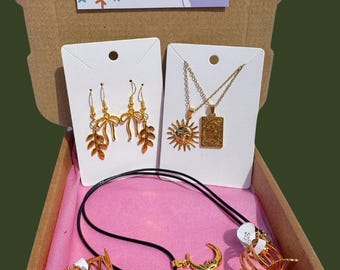 May include: A jewelry box with gold-colored earrings, necklaces, and rings. The earrings have a bow and leaf design. The necklaces feature sun and rectangular pendant designs. The rings have various geometric and floral patterns. The box has the text "Sanatorcrystals's JEWELLERY SHOP".