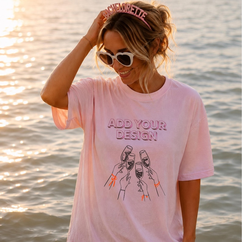 May include: A light pink short-sleeved t-shirt featuring the text ADD YOUR DESIGN in a bold font above a black line art graphic of four hands holding champagne flutes. The shirt is paired with a pink headband that reads BACHELORETTE in glitter.