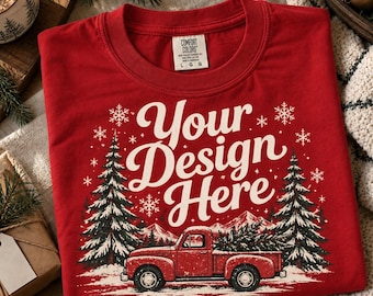Puede incluir: Una camiseta roja con un estampado gráfico de una camioneta roja antigua que transporta un pino, flanqueada por dos pinos grandes y copos de nieve. El texto "Your Design Here" está impreso en letra cursiva blanca sobre la camioneta.
