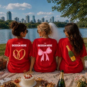 May include: Three red t-shirts with white text reading YOUR DESIGN HERE. The left shirt features a gold ring graphic, the center shirt has a pink bow, and the right shirt displays a green champagne bottle. The shirts are worn by three individuals.