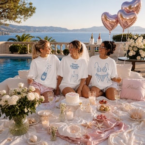 May include: Three white t-shirts featuring blue line art graphics of a champagne bottle, a hand gesture, and a striped bikini. The shirts are worn by three individuals sitting on a white blanket with a cake, flowers, and heart-shaped balloons.