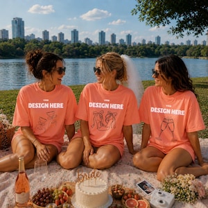 May include: Three coral-colored t-shirts featuring the white text YOUR DESIGN HERE and black line art illustrations of a gift, high-heeled shoes, and champagne flutes. The shirts are worn by three individuals sitting on a blanket outdoors.
